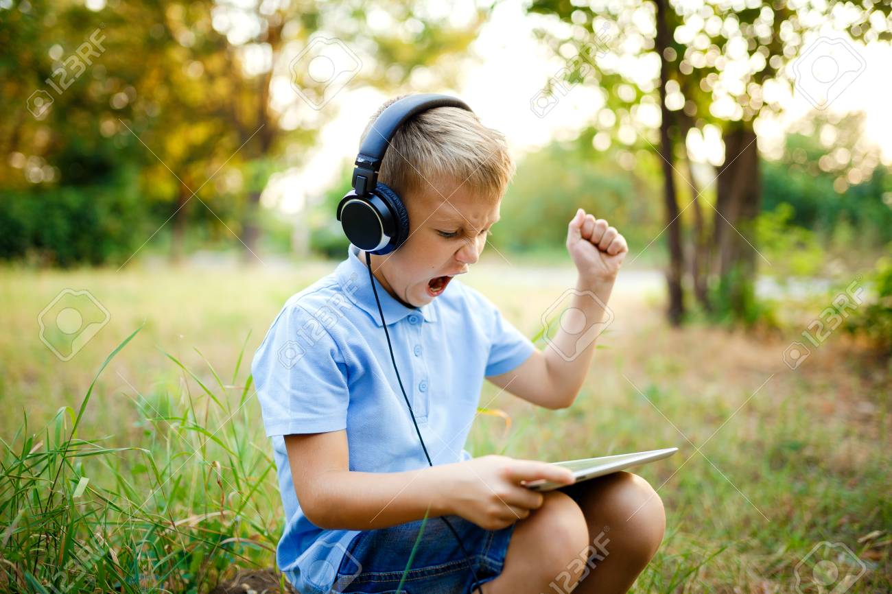 Young kid sitting in the forest wearing headphones and playing a game on tablet